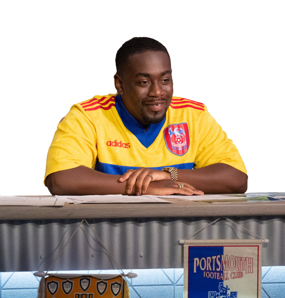 Andrew Mensah sitting behind a desk wearing a yellow football shirt 